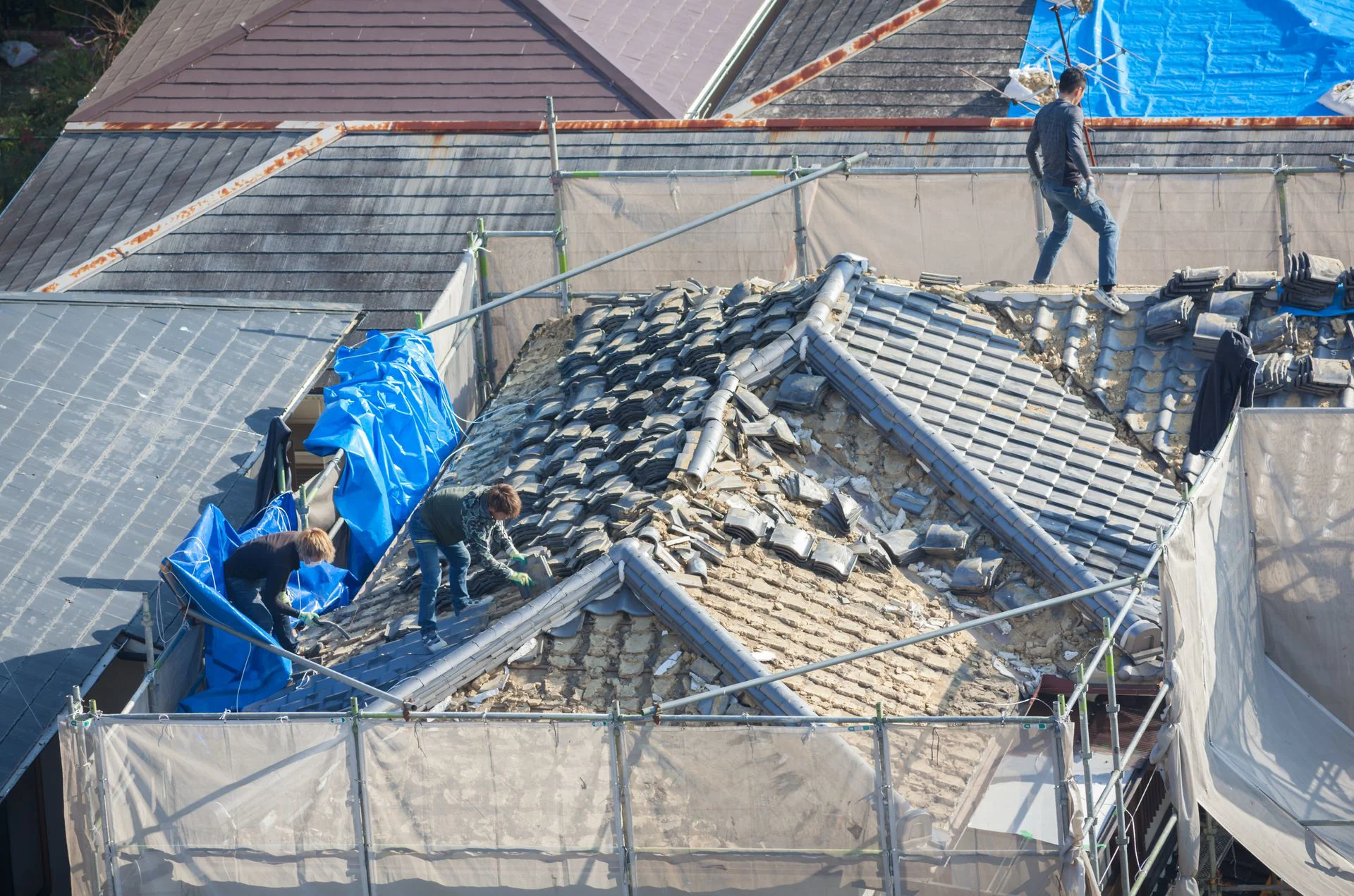 Workers are actively repairing a roof covered in broken tiles, with scaffolding and blue tarps surrounding the work area
